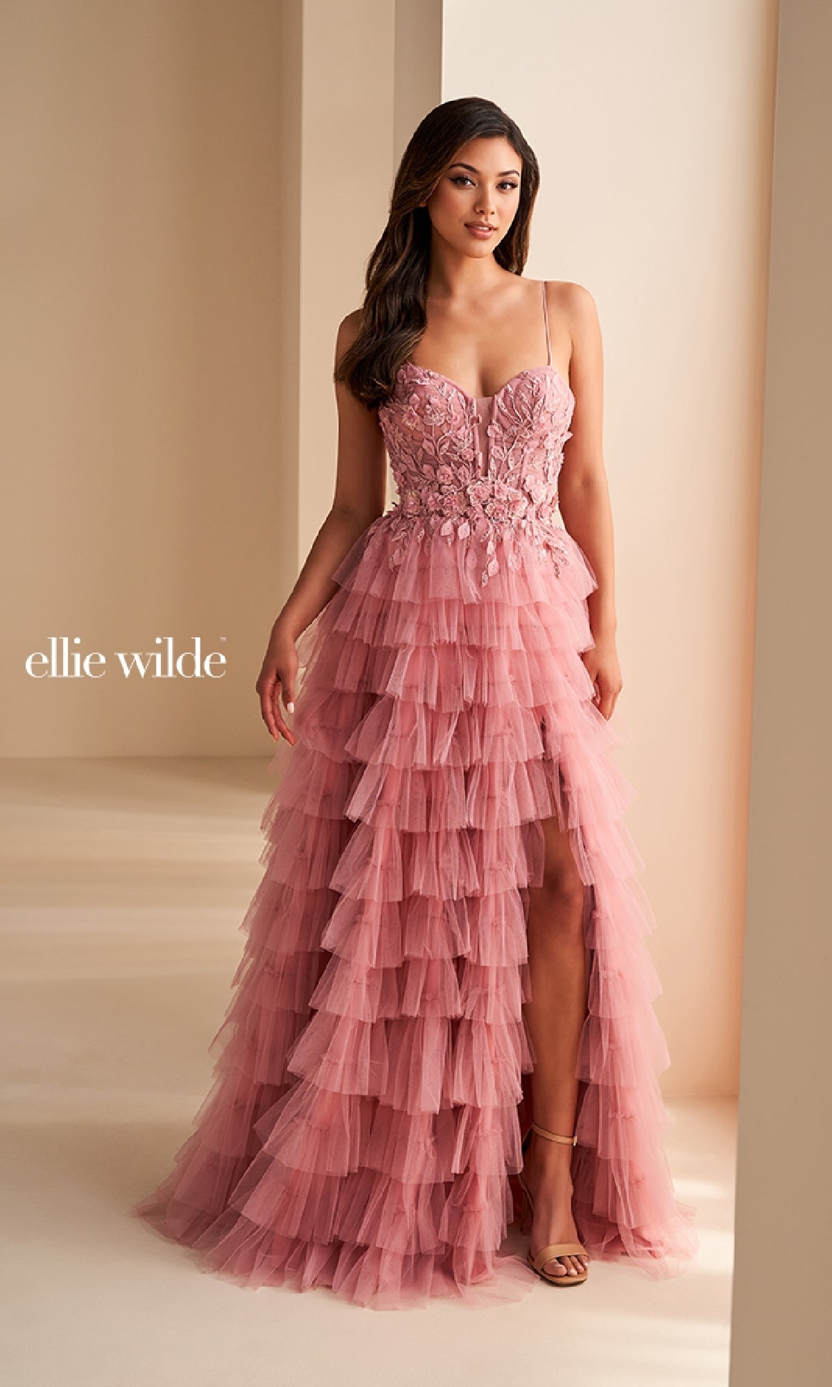 A woman stands in a room wearing the Ellie Wilde EW36223 long prom dress, featuring pink tiered ruffles, floral embroidery, spaghetti straps, and a thigh-high slit.
